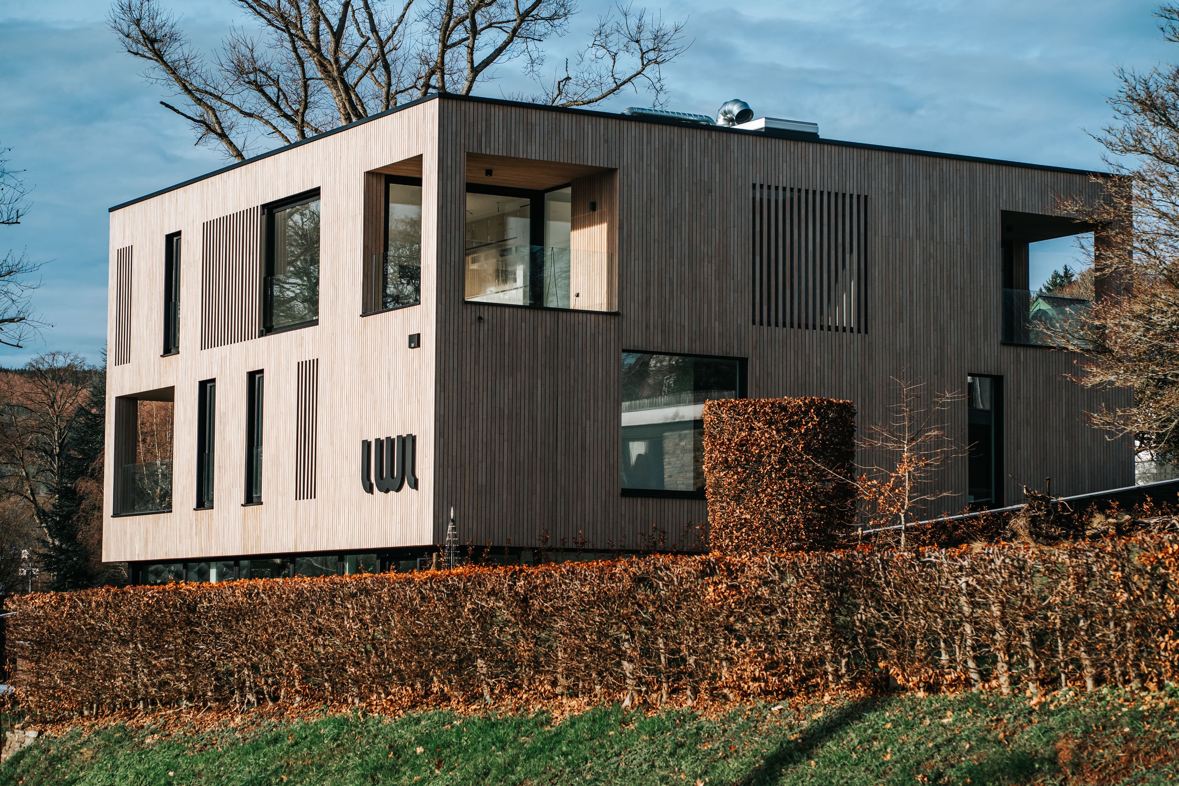 Bâtiment moderne en bois avec grandes fenêtres, entouré de haies, sous un ciel partiellement nuageux.