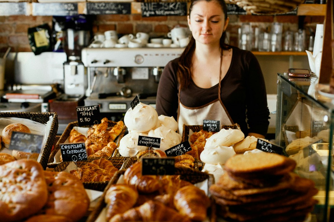 Une employée dans une boulangerie, entourée de pâtisseries variées, avec un comptoir en bois et des étiquettes de prix.