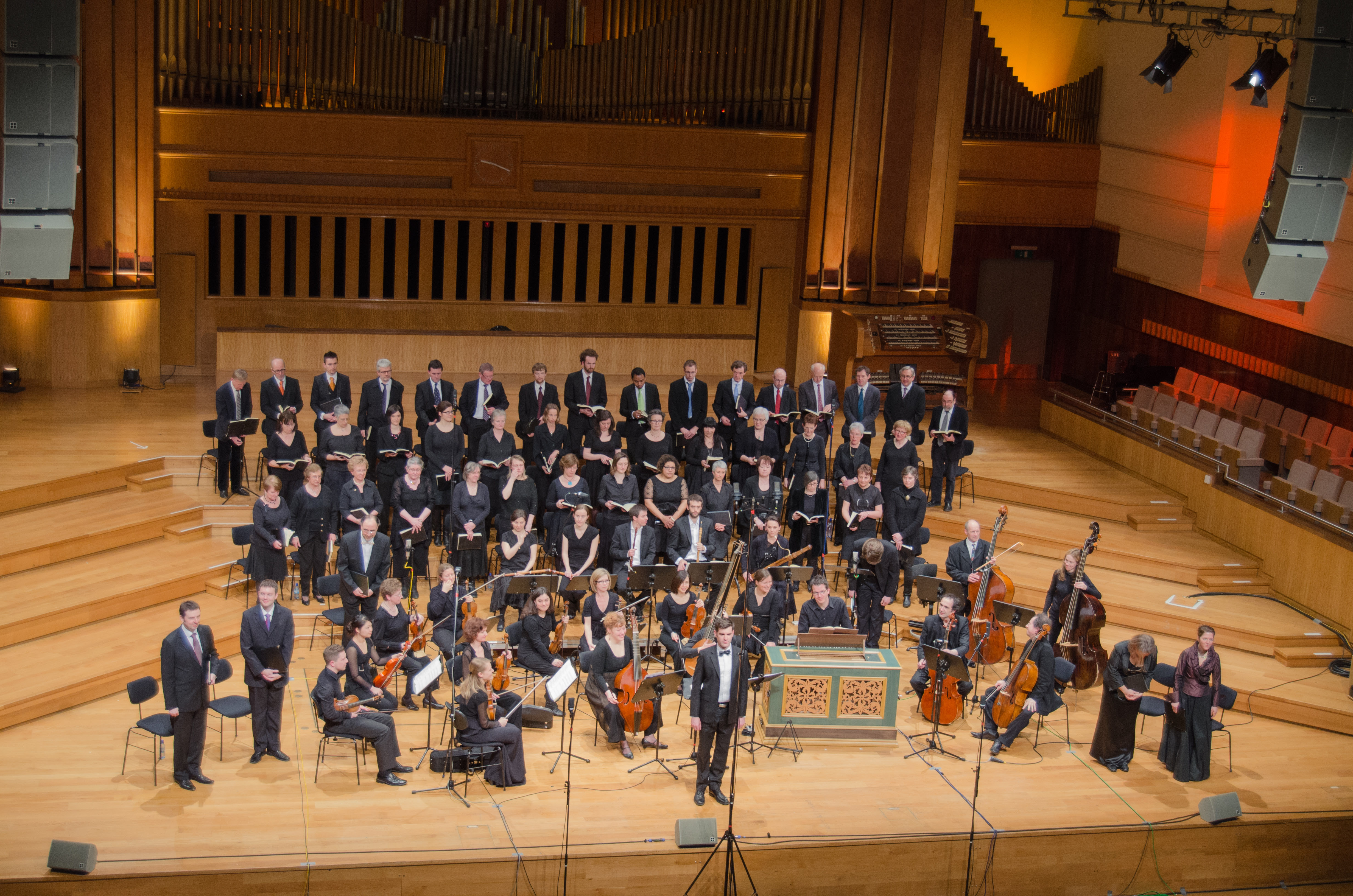 Een grote groep zangers en musici staat op een podium in een concertzaal, klaar voor een uitvoering van barokmuziek.