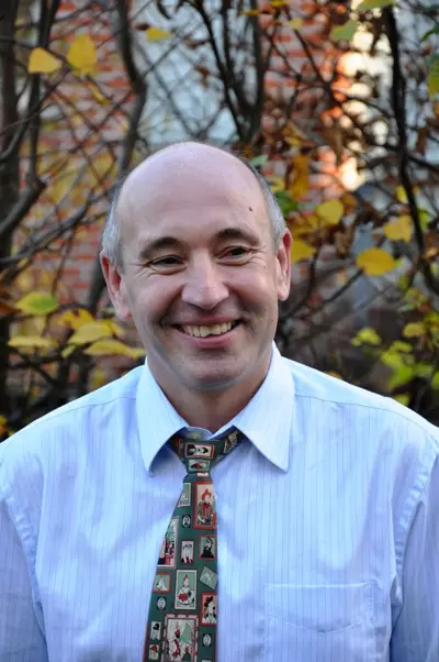 Un homme souriant portant une chemise claire et une cravate colorée, entouré d'un environnement naturel avec des feuilles jaunes.