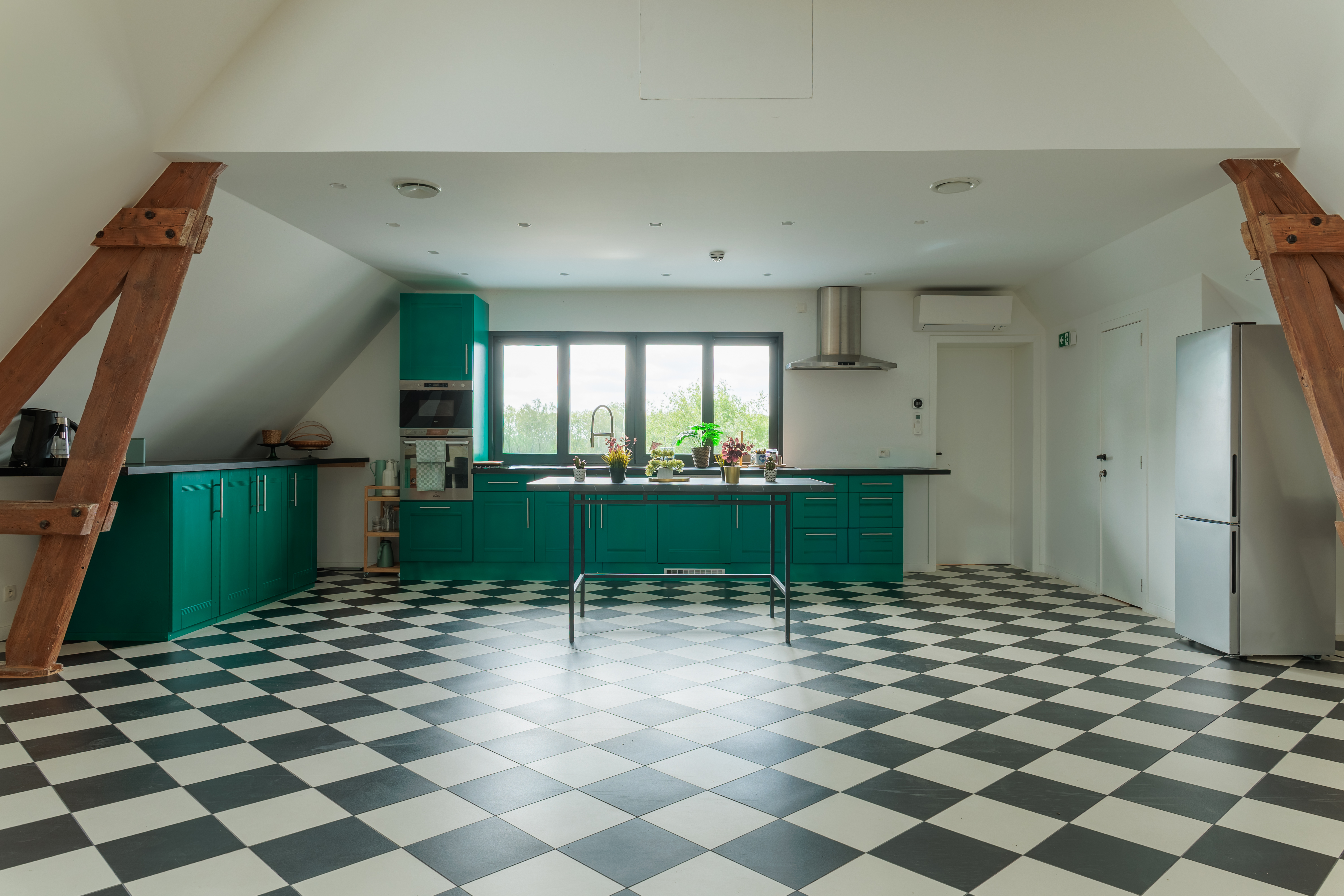 Cuisine spacieuse avec un sol en damier noir et blanc et des meubles de cuisine verts, baignée de lumière naturelle.