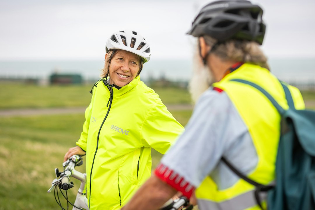 Deux cyclistes souriants en vêtements de sécurité lumineux, stand sur un chemin vert face à un paysage naturel.