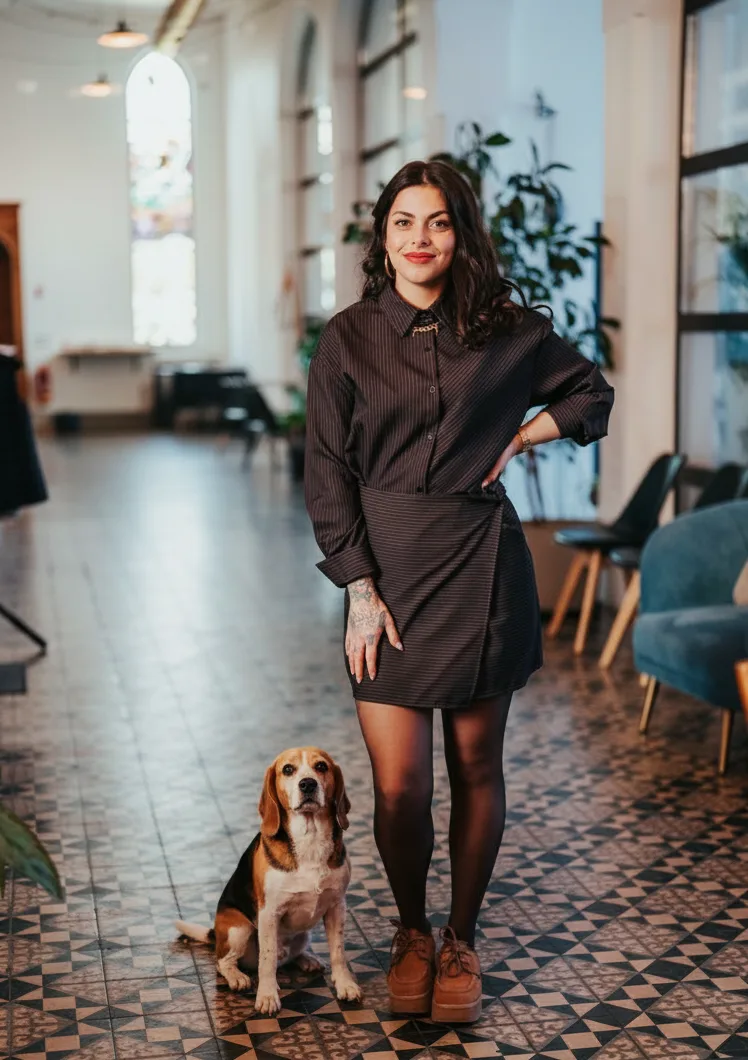 Une femme souriante avec une tenue noire se tient dans un espace lumineux, aux murs blancs, accompagnée d'un chien Beagle.