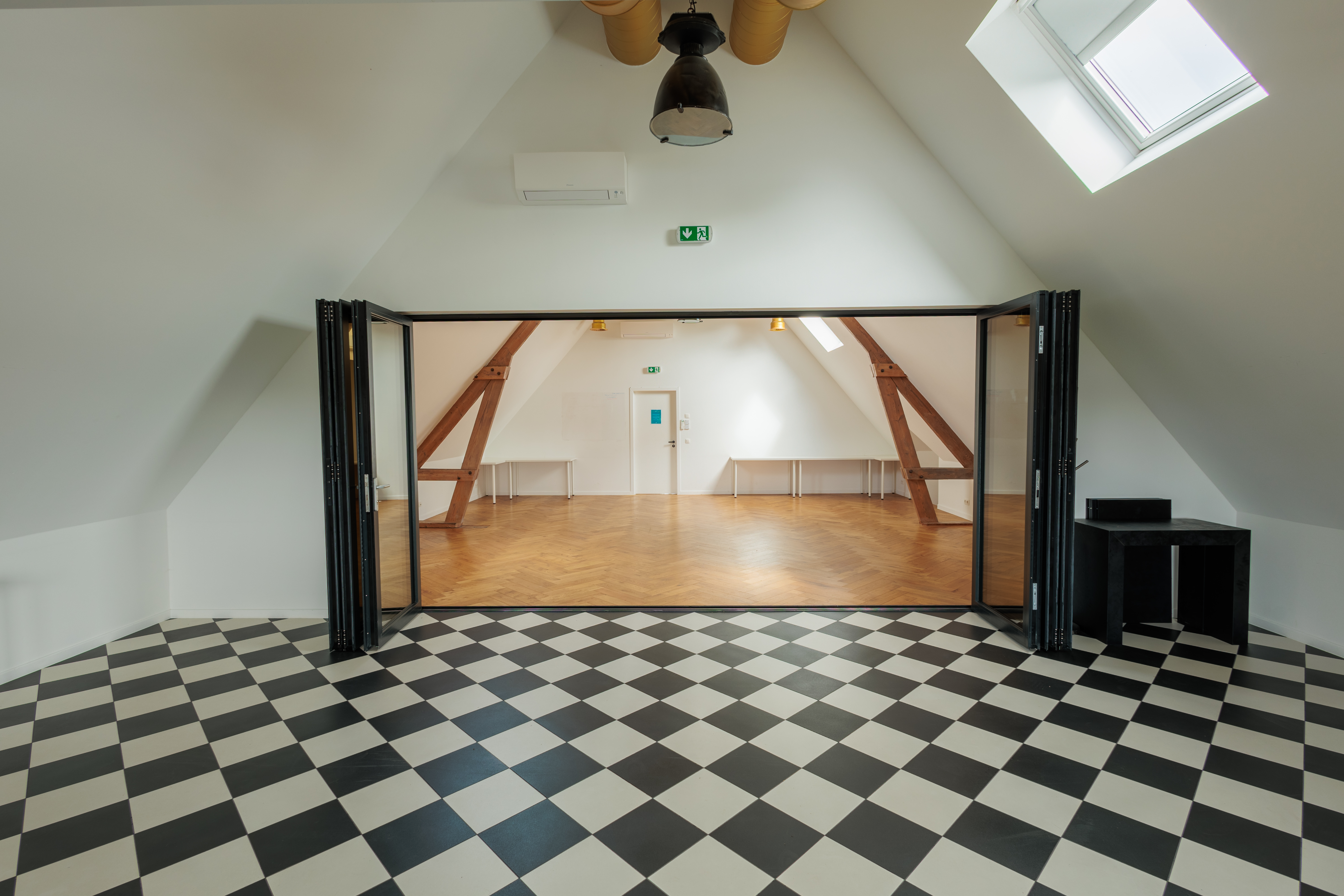 Espace événementiel lumineux avec un sol en damier noir et blanc, murs blancs et poutres apparentes, ambiance chaleureuse.