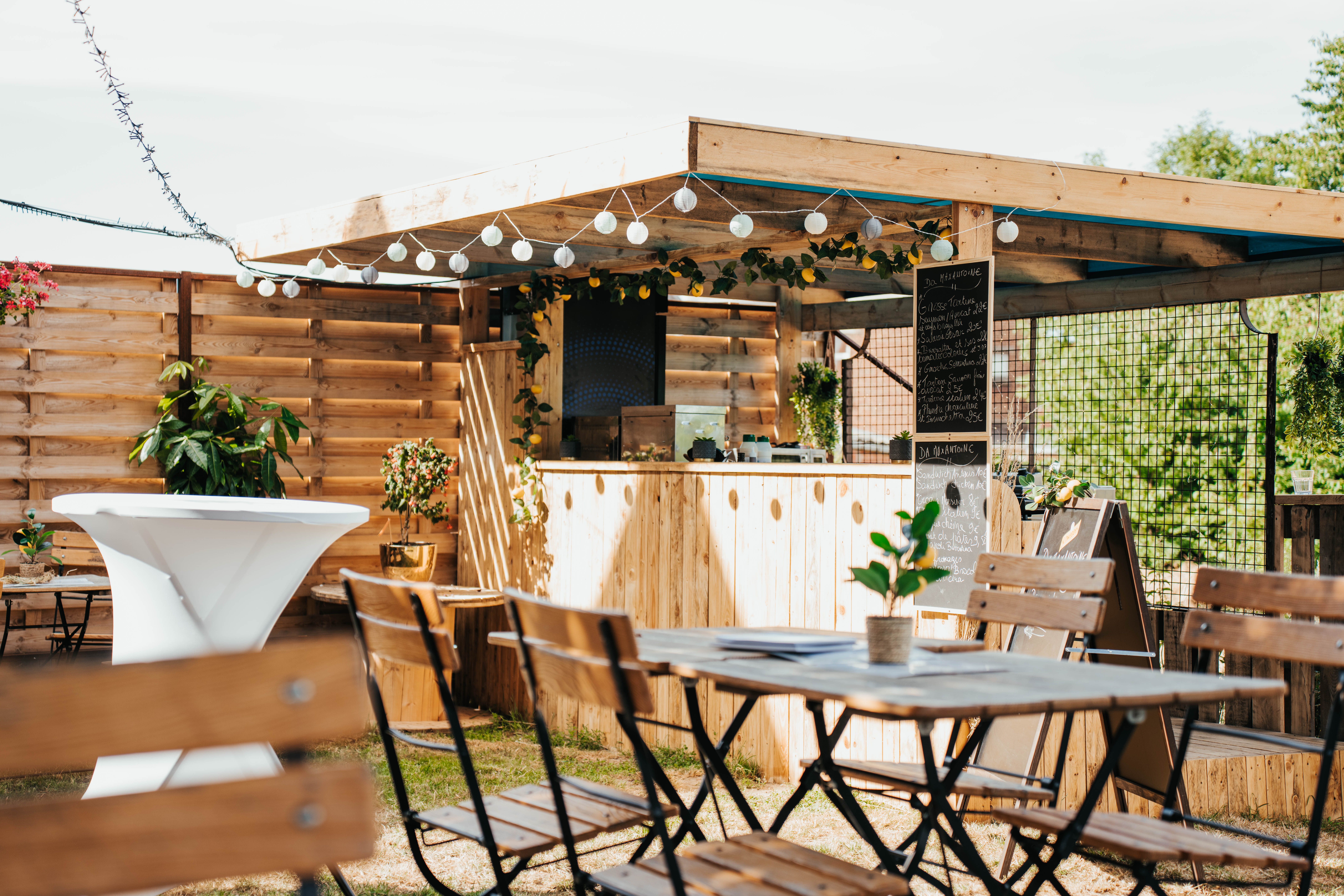 Espace extérieur convivial avec tables en bois et un bar, illuminé par des guirlandes lumineuses, créant une ambiance chaleureuse.