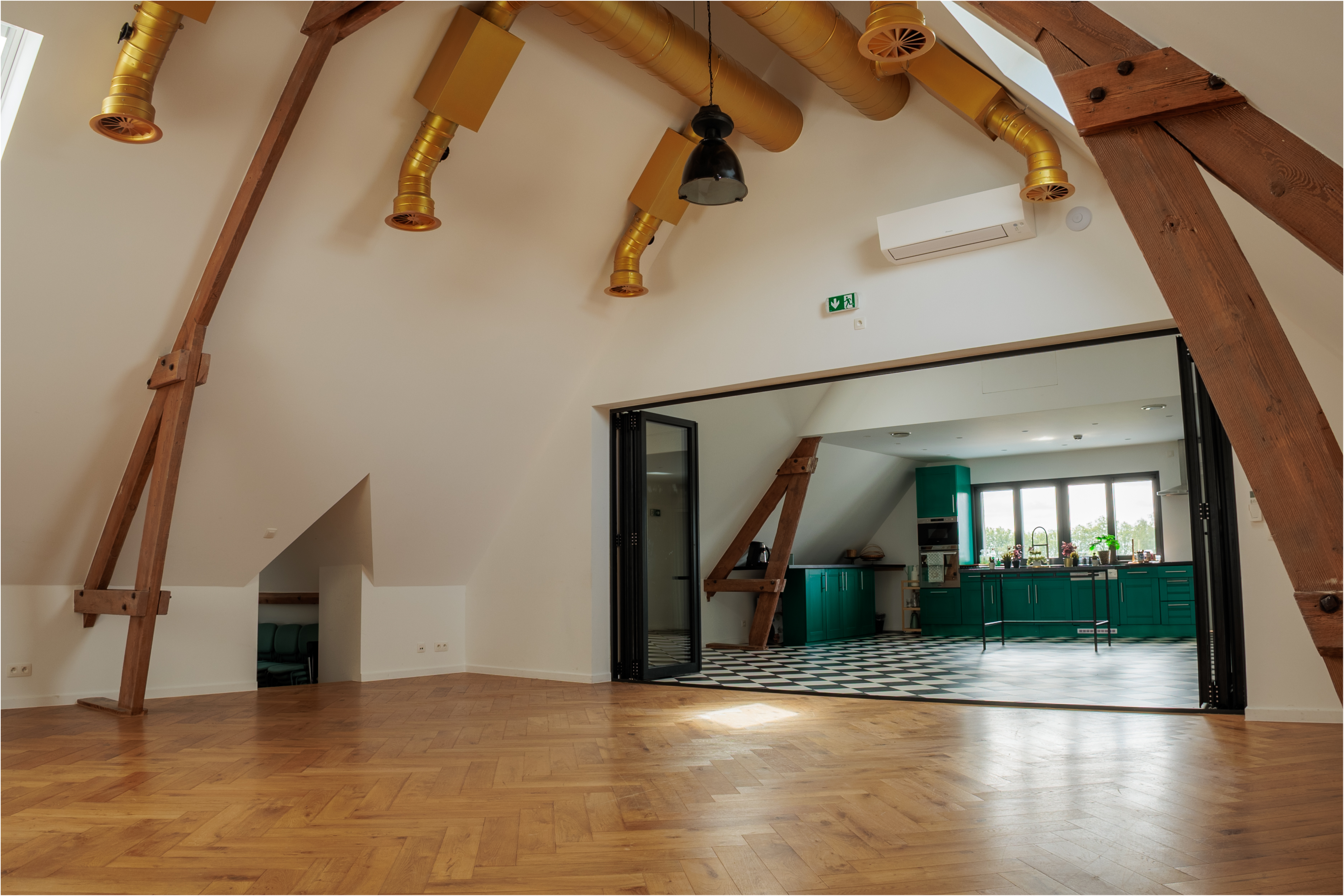 Un espace intérieur moderne avec des poutres apparentes, un sol en bois et une cuisine colorée, baigné de lumière naturelle.