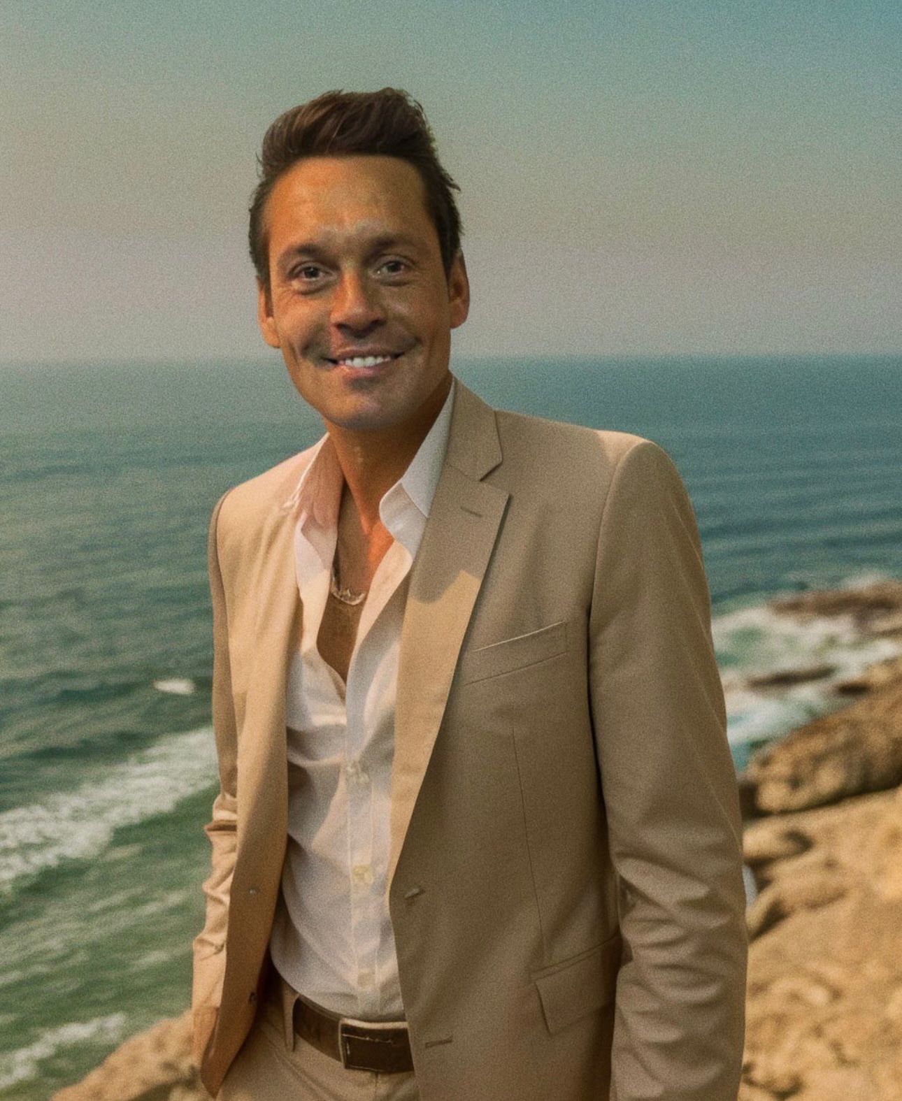 Un homme souriant en costume clair se tient devant la mer, avec des vagues en arrière-plan et un ciel ensoleillé.