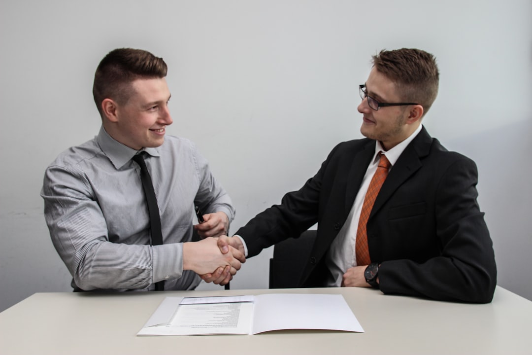 Deux hommes en costume se serrent la main autour d'une table avec des documents, ambiance professionnelle, lumière naturelle.