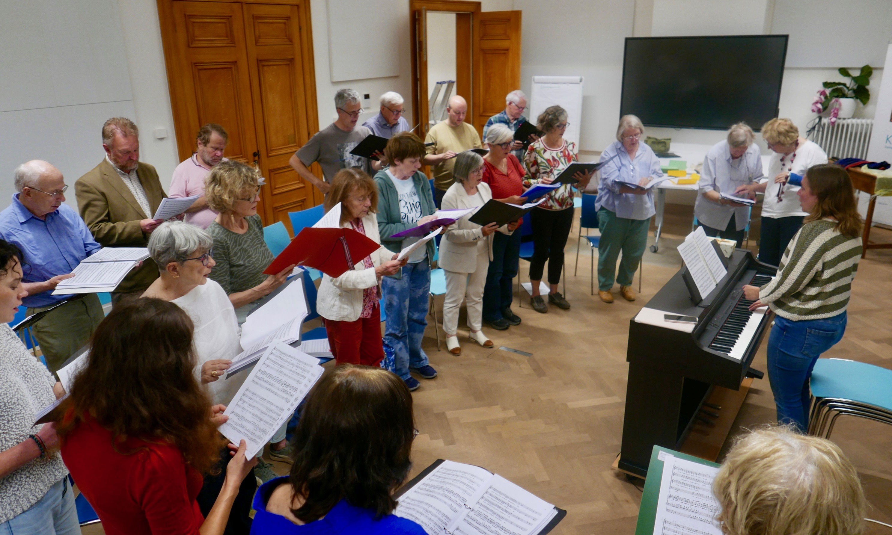 Een groep zangers repeteert in een lichte zaal met partituren en een piano. De setting is gezellig en ontspannen.