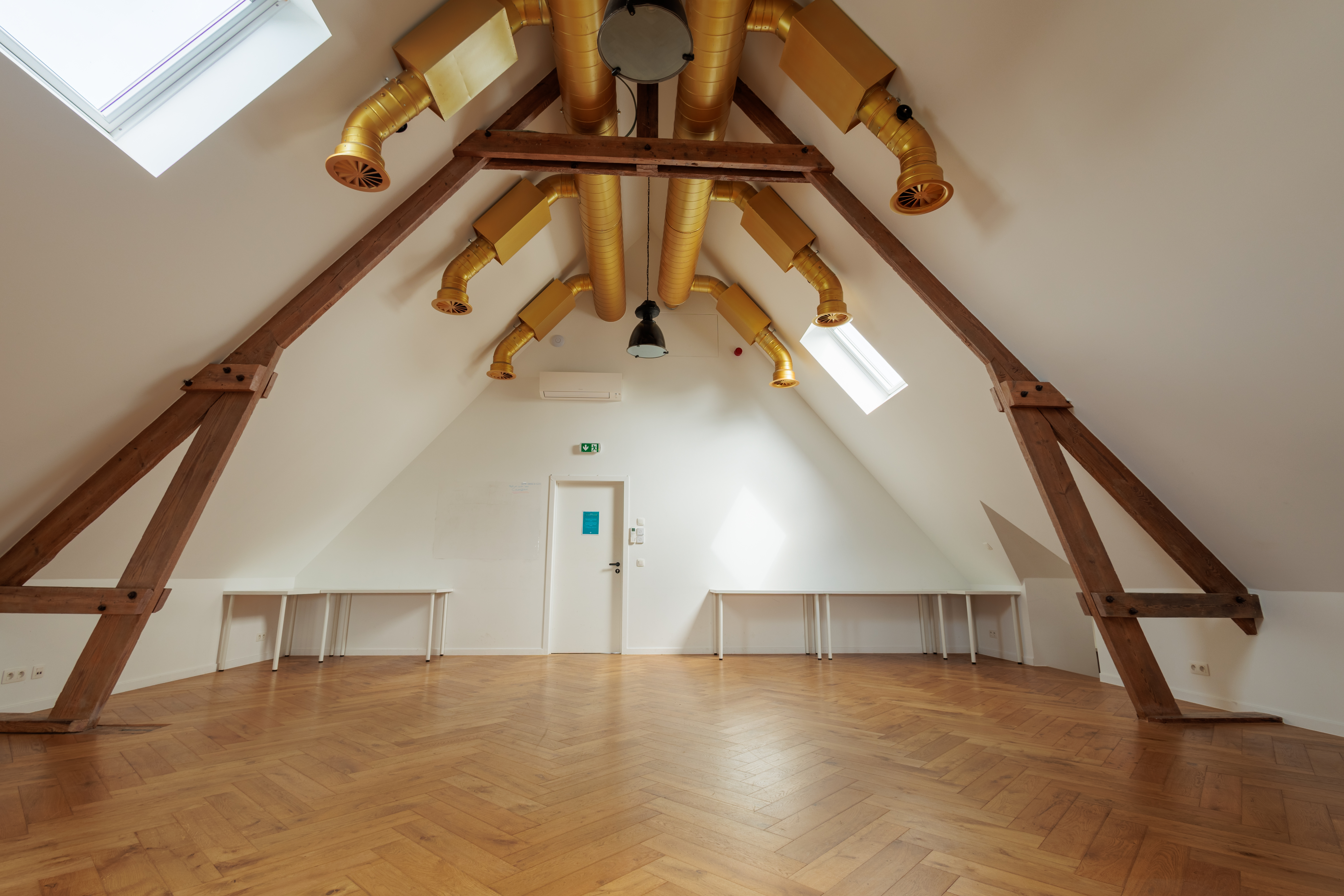Salle lumineuse avec poutres en bois, fenêtres de toit et système de ventilation doré, offrant un espace accueillant.