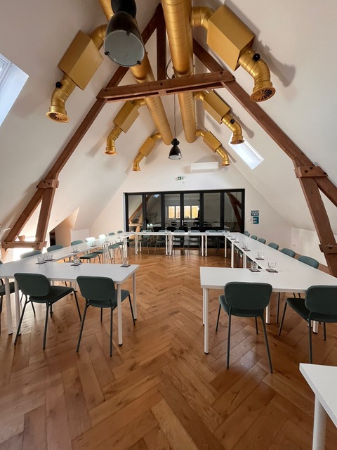 Salle de réunion lumineuse avec des tables blanches et des chaises vertes, poutres en bois apparentes et fenêtres offrant une vue extérieure.