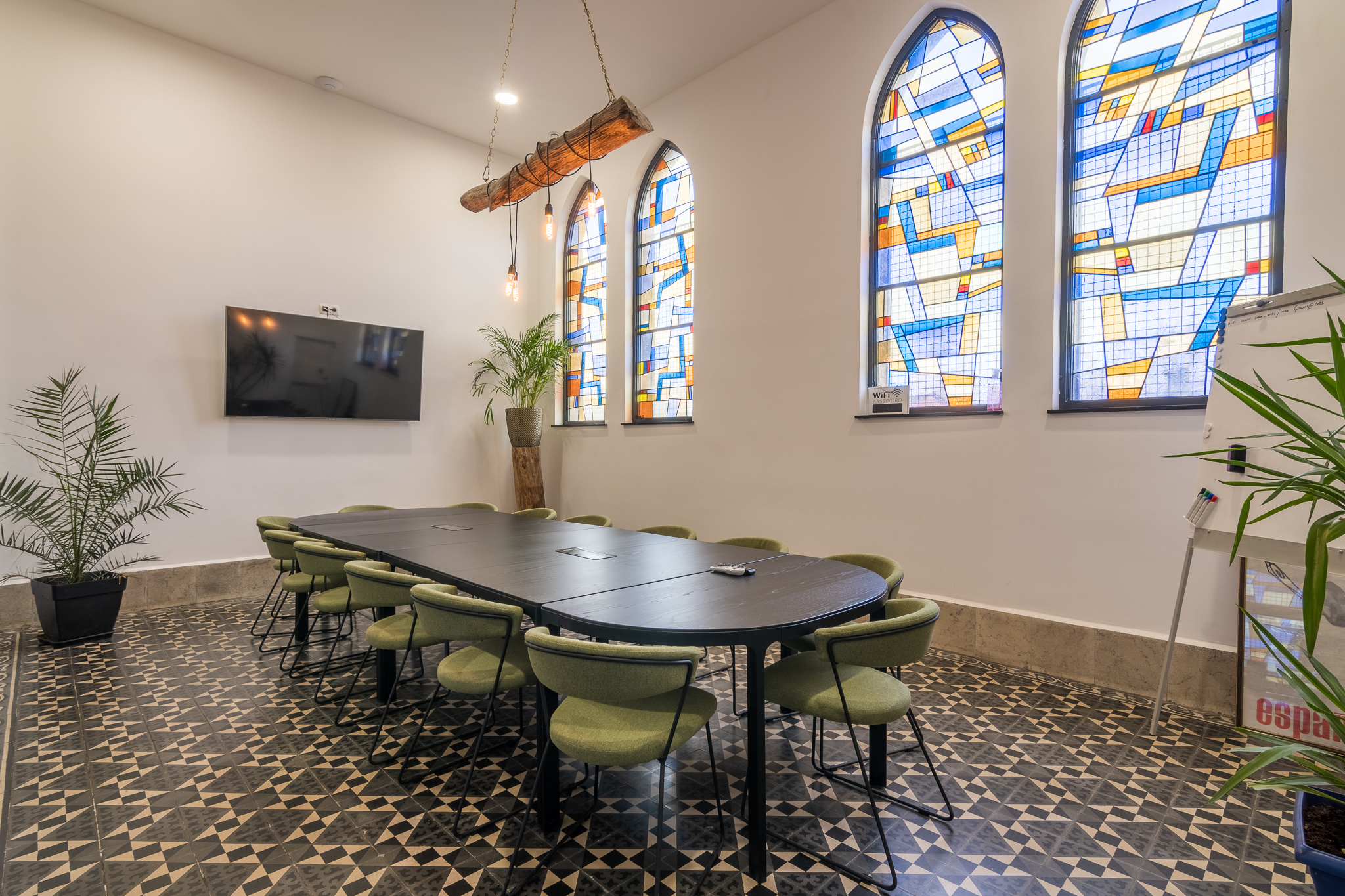 Salle de réunion spacieuse avec grande table, chaises vertes et vitraux colorés, ambiance chaleureuse et professionnelle.