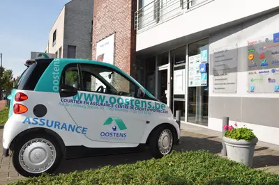 Une petite voiture de marque Smart avec l'inscription Oostens Insurance stationnée devant le bureau d'assurance à Wemmel, sous un ciel ensoleillé.