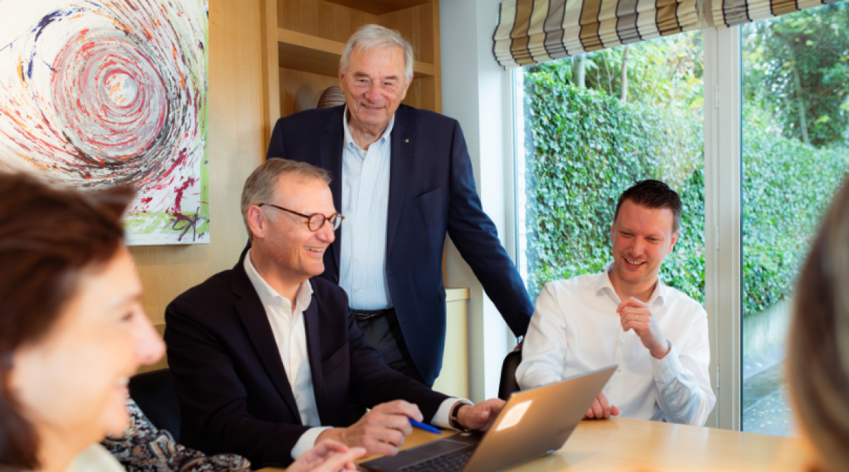 Un groupe de personnes souriantes se rencontre autour d'une table, avec un ordinateur portable ouvert, dans un espace lumineux avec des fenêtres et des œuvres d'art accrochées au mur.