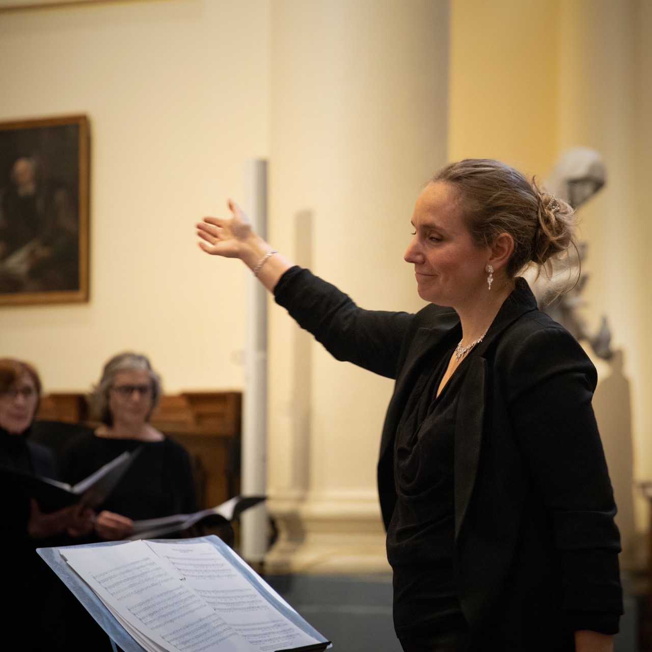 Une cheffe de chœur dirige un ensemble vocal dans une salle lumineuse, avec des partitions visibles au premier plan.