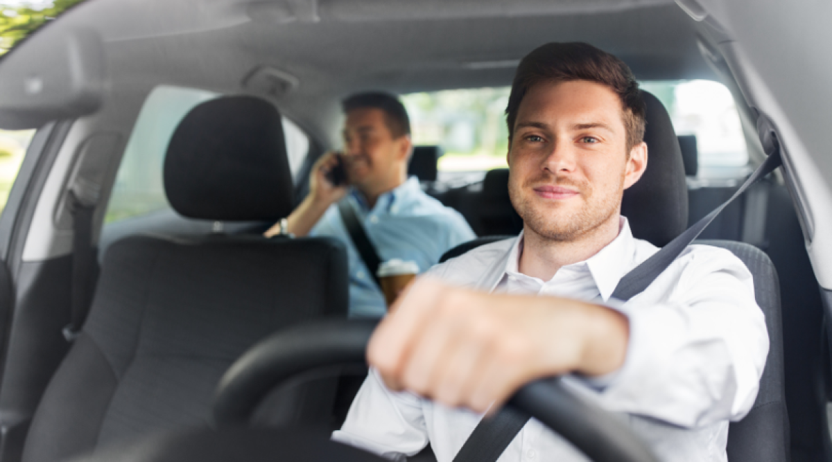 Un homme souriant conduit une voiture tandis qu'un passager à l'arrière parle au téléphone.
