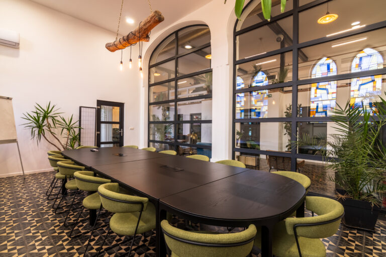 Salle de réunion moderne avec une grande table noire entourée de chaises vertes, des murs avec de grandes fenêtres et des éléments décoratifs en plantes.