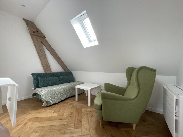 Un salon minimaliste sous les combles avec un canapé vert, un fauteuil vert, une table blanche et des poutres apparentes.