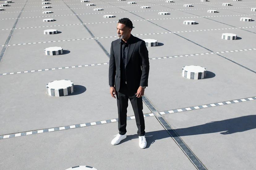 Un homme en costume noir et baskets blanches se tient debout sur un sol moderne avec des éléments circulaires et des lignes blanches marquées.