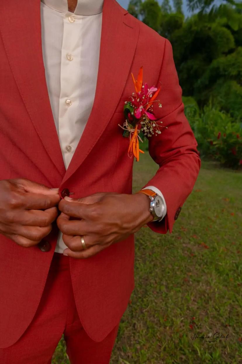 Un homme en costume rouge boutonne sa veste, avec une boutonnière colorée et une montre apparente, dans un décor verdoyant.