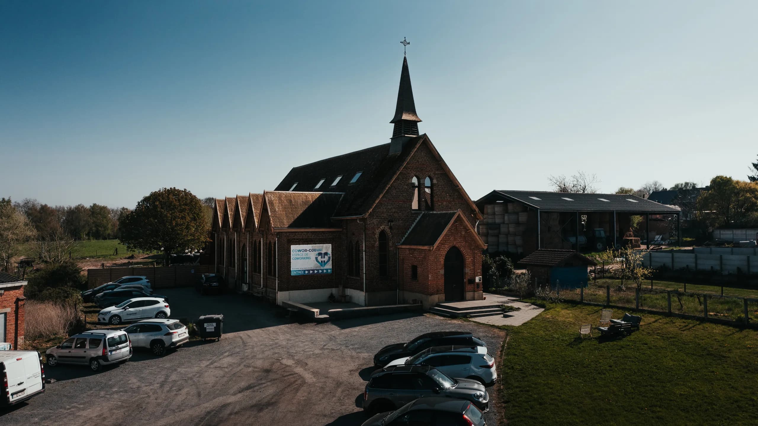 Une église en briques avec un clocheton, entourée de voitures stationnées et de champs verdoyants.