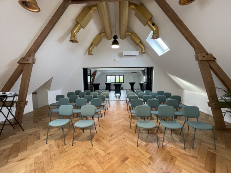 Une salle de réunion avec des chaises bleu-vert disposées en rangées, des poutres en bois, et des conduits d’aération dorés.
